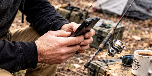 Angler using Pine 9 JettyHawk fishing rod holster to secure spinning rod while checking smartphone at shoreline campsite preventing rod from being set on ground while managing gear and preparing tackle