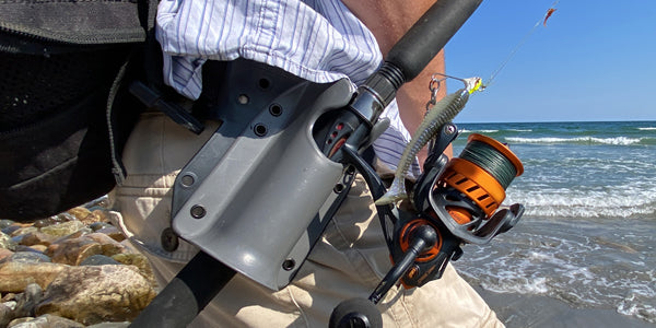 Pine 9 JettyHawk fishing rod holster secured to belt during surf fishing holding spinning rod above rocks and sand preventing reel damage while angler changes lure and manages gear near ocean shoreline