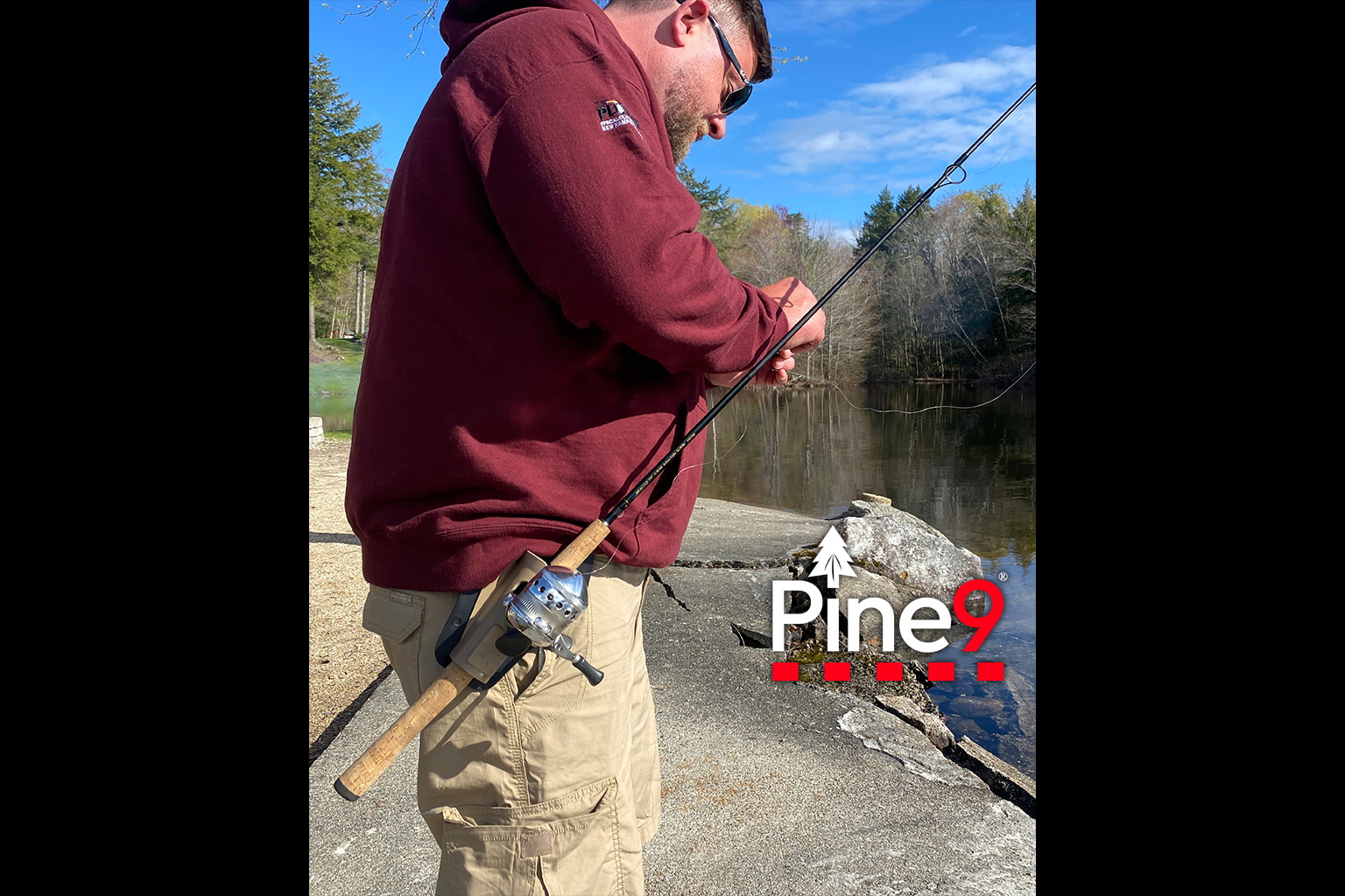 pine 9 JettyHawk fishing rod holster hands free hip rod hip holder man with maroon hoodie fixes his fishing lure next to a lake with his rod in his hip holder
